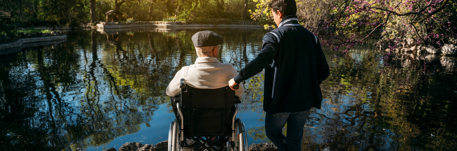 Ein junger Mann und ein alter Mann auf dem Rollstuhl vor einem kleinen See